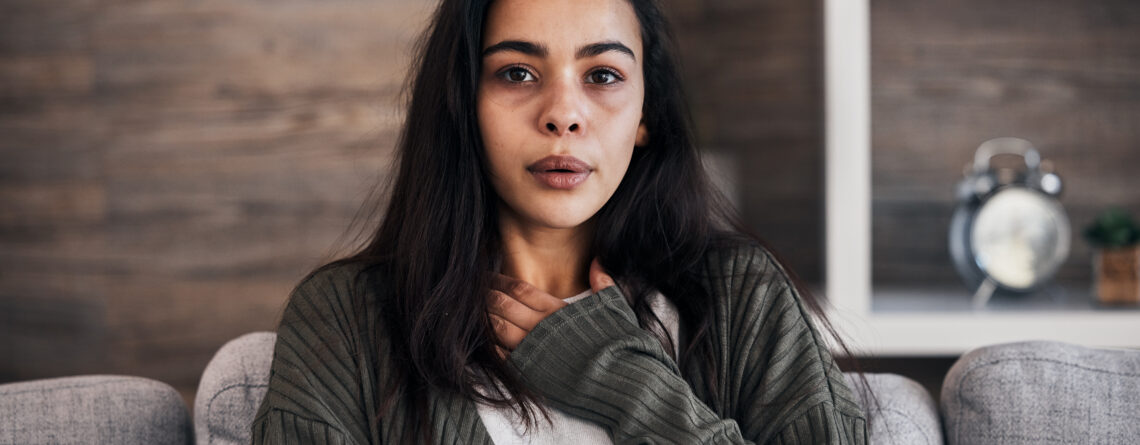 Person sitting on a couch with one hand on their chest, experiencing one of the trauma responses. Background includes wooden wall and shelf with an alarm clock.