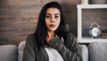 Person sitting on a couch with one hand on their chest, experiencing one of the trauma responses. Background includes wooden wall and shelf with an alarm clock.