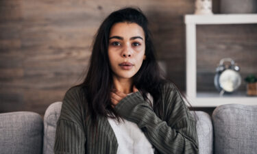 Person sitting on a couch with one hand on their chest, experiencing one of the trauma responses. Background includes wooden wall and shelf with an alarm clock.