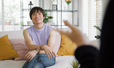 A non-binary Asian individual sits comfortably on a couch during a face-to-face session with an LGBTQ+ affirming therapist, in a bright, welcoming room with natural light and homey decor.