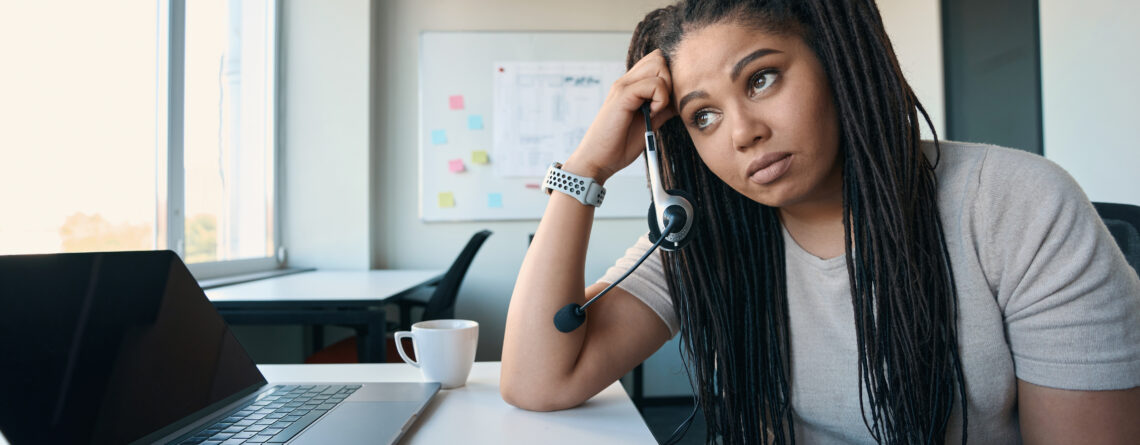 Tired female office worker with ADHD symptoms headset in hand leaning on desk and staring into distance