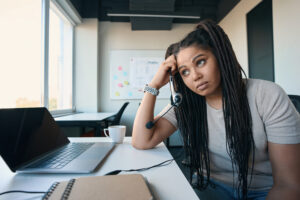 Tired female office worker with ADHD symptoms headset in hand leaning on desk and staring into distance