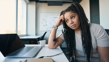 Tired female office worker with ADHD symptoms headset in hand leaning on desk and staring into distance