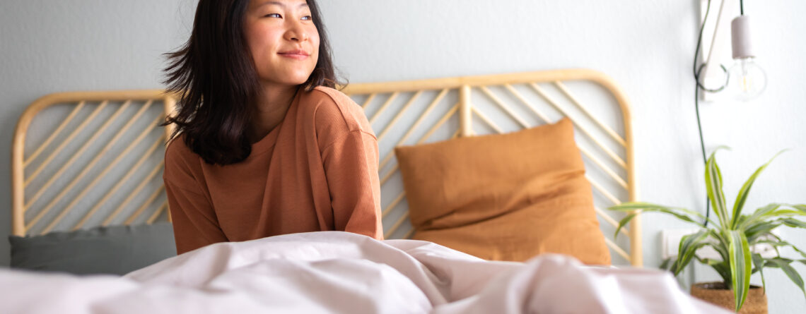 Happy young asian woman sitting on bed after waking up in the morning in beautiful, cozy bedroom at home after CBT-I treatment