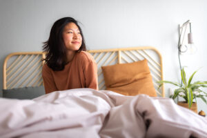 Happy young asian woman sitting on bed after waking up in the morning in beautiful, cozy bedroom at home after CBT-I treatment