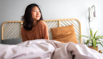 Happy young asian woman sitting on bed after waking up in the morning in beautiful, cozy bedroom at home after CBT-I treatment